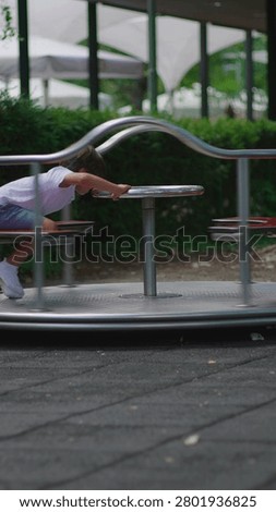 Young boy rotating roundabout while standing upright and gripping steering wheel expressing coordination energy and active engagement within lively playground environment