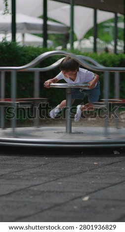 Young boy rotating roundabout while standing upright and gripping steering wheel expressing coordination energy and active engagement within lively playground environment