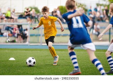 Young Boy Running Fast And Kicking Soccer Ball On Grass Pitch. Football Tournament For Youth School Teams. Group Of Children In Jersey Unifrom Playing Sports. Football Fans And Parents In Background