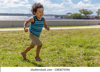 A Young Boy Runs Across The Field While Laughing Out Loud. The Happy Toddler Is Having A Wonderful Time Running Outdoors In The Waterfront Park.