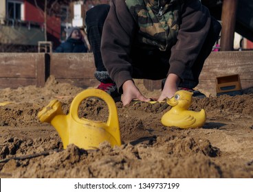 Young Boy In The Sandbox. Children Is Playing Outdoor In A Sandpit. Toys – Yellow Plastic Duck And Teapot In The Sand Pit