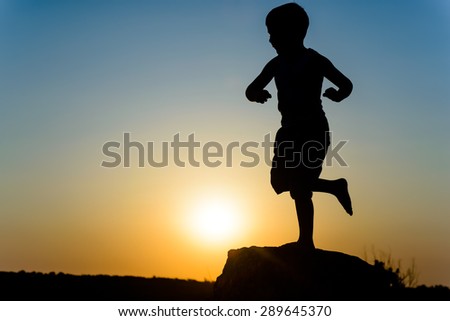 Young boy silhouetted by the sunset standing balancing on one leg against the fiery orange orb of the sun in a twilight sky