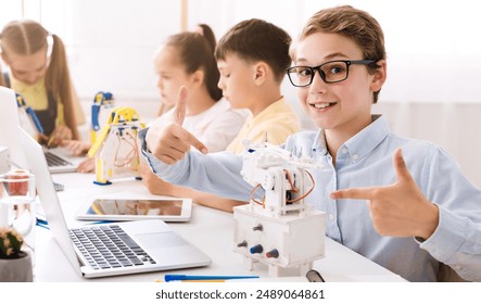 A young boy sits in a classroom setting with other students. He is smiling and pointing at a small robot he has built, demonstrating his learning and enthusiasm for STEM education. - Powered by Shutterstock