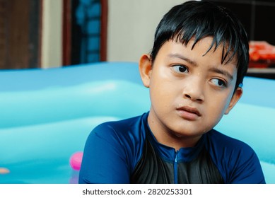 Young boy sits in inflatable pool, eyes wide, expression curious. Water droplets cling to his dark, damp hair. He wears blue swim top with black panel. Background shows blurred pool and wooden doorway