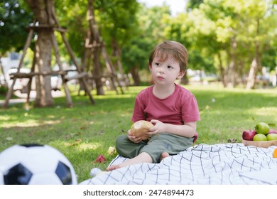 Young boy sits on blanket, holding bread, picnic basket with fruits nearby, soccer ball, outdoor family picnic scene. Relaxing family vacation Caucasian people on the weekend - Powered by Shutterstock