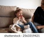 A young boy sits on a couch, biting into a red apple with a pained expression. He is accompanied by his Algerian mother, creating a cozy indoor atmosphere.