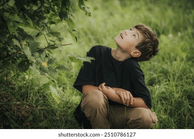 A young boy sits on the grass, gazing up at the tree branches above him with curiosity and wonder. The lush green surroundings and his thoughtful expression create a peaceful, nature-focused scene. - Powered by Shutterstock