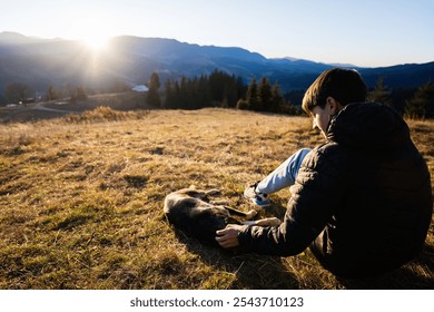 A young boy sits on a grassy field, gently petting his dog while watching a serene sunset over the mountains. The setting conveys tranquility and the beauty of nature. - Powered by Shutterstock