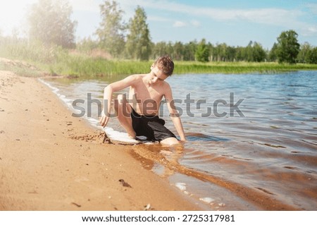 Young boy sits on the sandy lakeside, focused and creative, building a sandcastle under a clear blue sky surrounded by lush green trees, enjoying a warm, sunny day.
