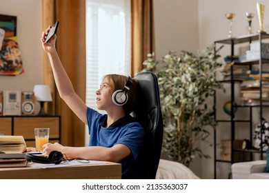 Young Boy Sits In A Room At Desk Has Remote Teaching Internet Lessons. Child Has Lost Coverage On Phone, Poor Internet Connection, Pulls Smartphone Up In Search Of A Better Network.