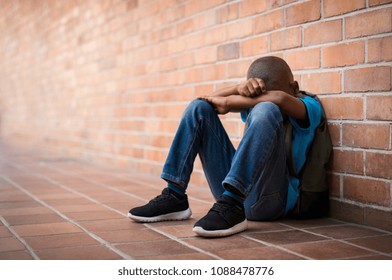 Young boy sitting alone with sad feeling at school. Depressed african child abandoned in a corridor and leaning against brick wall. Bullying, discrimination and racism concept with copy space. - Shutterstock ID 1088478776