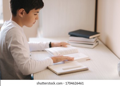 Young Boy Sitting At Desk Read The Book And Whright Down In Notebook. Study At Home During Quarantine