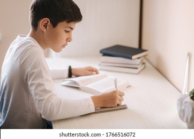 Young Boy Sitting At Desk Read The Book And Write Down In Notebook. Study At Home During Quarantine