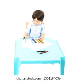 Young Boy Sitting Down At A Table With Paper And Crayons Against A White Background