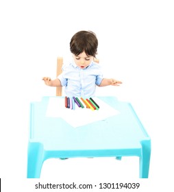 Young Boy Sitting Down At A Table With Paper And Crayons Against A White Background