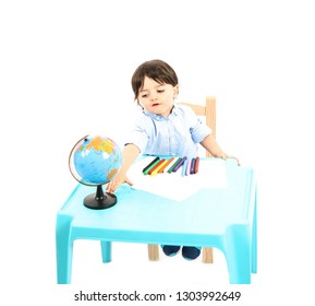 Young Boy Sitting Down At A Table With Paper And Crayons Against A White Background