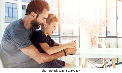 Young boy sitting with father at the table and playing together pc tablet in modern loft. Horizontal, blurred background. Sunlights effect