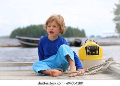 A Young Boy Is Sitting On A Dock Next To A Minnow Bucket, Ready To Go Fishing. He Has A Rope Around His Foot. There Is A Boat In The Background.