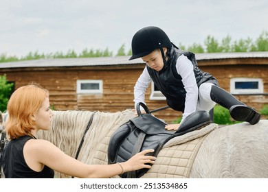 Young boy sitting on horse while woman helping him adjust saddle during horse riding training session in countryside, wooden cabin in background - Powered by Shutterstock