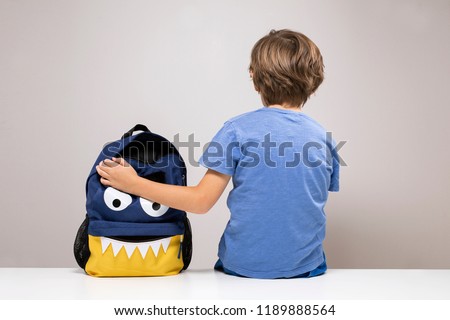 Young boy sitting on white desk with his cute school backpack, monster face bag with open pocket. Back to school