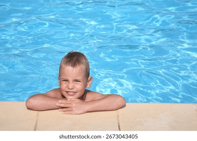 A young boy is smiling and looking at the camera while sitting in a pool. The water is clear and calm, and the boy appears to be enjoying his time in the pool