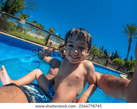 Young boy smiling while floating in a pool on a sunny day at a water park, surrounded by vibrant greenery and clear blue skies
