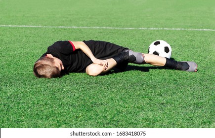 A Young Boy Soccer Player Injured His Leg During The Match. Sufferer Child Football Player And Ball On The Field. Kids Injury In Sport Concept. Children's Football School. Background. Copy Space