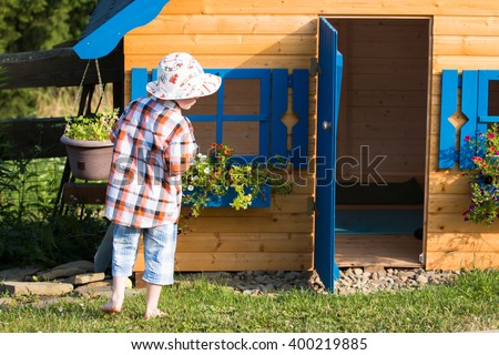 young boy standing and playing outside the children wooden playhouse with blue windows and blooming flowers in the countryside garden