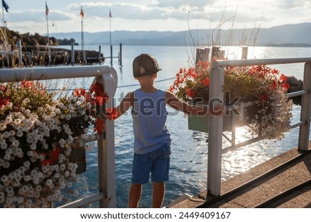A young boy stands on a pier in a small village on Lake Leman, surrounded by ornate flower boxes, enjoying the tranquil scenery and the gentle touch of the sunset on the water.