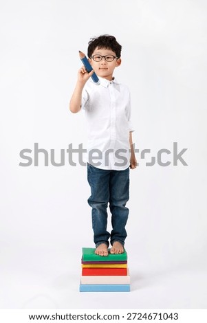 Young boy stands on stack of books holding large pencil in his hand while wearing glasses and casual clothes in a simple white background