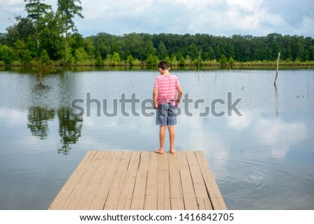 A young boy stands on a wooden fishing pier that sits on a freshwater bass pond and looks down toward the water on a hot summer day in July located in Louisiana.