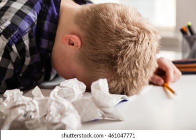 Young boy struggling with his homework sitting with his head down on the table surrounded by crumpled pages of screwed up paper