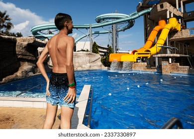 Young boy in swimming trunks and shorts looking at water slides in aquapark.