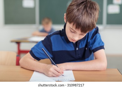 Young boy taking notes in class at school bending over his paperwork as he works, blackboard and student in the background