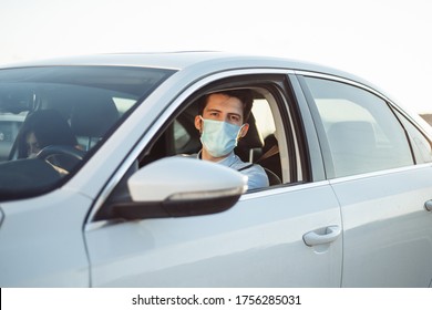 Young Boy Taxi Driver Gives Passenger A Ride Wearing Sterile Medical Mask. A Man In The Car Behind The Steering Wheel Works During Coronavirus Pandemic. Social Distance And Health Safety Concept.