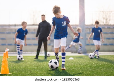 Young Boy Training Sports In Sunny Summer Day. Happy Kids Practicing Soccer On Schoolyard. Young Coach With Children Football Players On Training Session. Sports Stadium Seats In The Background