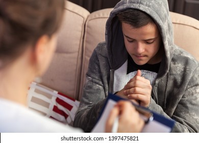 Young boy in trouble sitting on sofa attending a counseling session at the psychologist or social services professional, clasping hands, close up