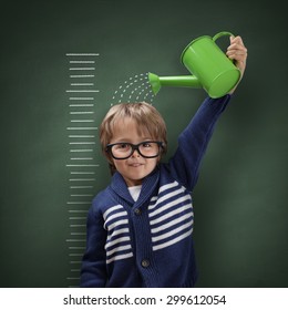 Young boy trying to make himself taller with watering can measuring his growth in height against a blackboard scale