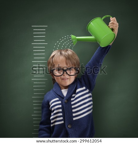Young boy trying to make himself taller with watering can measuring his growth in height against a blackboard scale