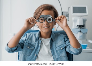 Young boy undergoing eye test with spectacles in medical clinic. Small patient at optician office choosing selecting eye sight lenses glasses with ophthalmic medical equipment - Powered by Shutterstock