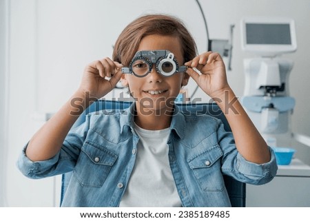 Young boy undergoing eye test with spectacles in medical clinic. Small patient at optician office choosing selecting eye sight lenses glasses with ophthalmic medical equipment
