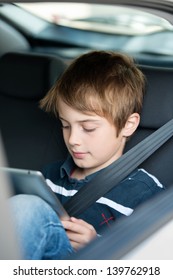 Young Boy Using A Tablet Computer While Sitting In The Back Passenger Seat Of A Car With A Safety Belt Over His Shoulder