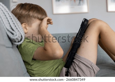 Young boy using tablet for online learning at home, sitting on couch and exploring educational apps or games, combining digital technology with early childhood development and study habits.