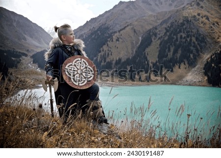 A young boy in the viking cosplay costume holding a wooden shield and a axe and standing next to the beautiful alpine lake with Mountains view