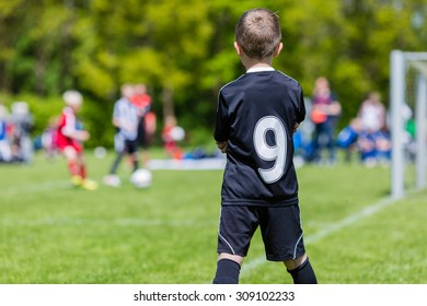 Young Boy Watching His Team Mates Play A Kids Soccer Match On Soccer Field With Green Grass.