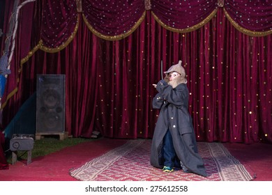 Young Boy Wearing Clown Make Up, Hat and Over Sized Coat and Holding Sword on Stage with Red Curtain