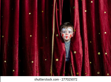 Young Boy Wearing Clown Make Up Peering Out Through Opening in Red Stage Curtains