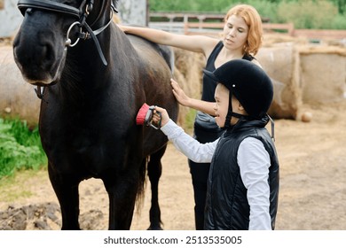 Young boy wearing helmet brushing horse under supervision of woman on farm. Both engaging in bonding activity, horse standing calmly while being groomed - Powered by Shutterstock