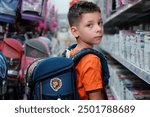 Young boy wearing an orange shirt tries on a new backpack in a store aisle, surrounded by various school supplies and bags