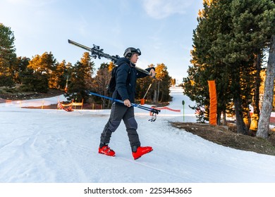 Young boy wearing ski clothes and carrying ski equipment on his shoulders. He is looking away and carrying the ski sticks on the left hand. Snow can be seen. Finishing the ski day coming home.